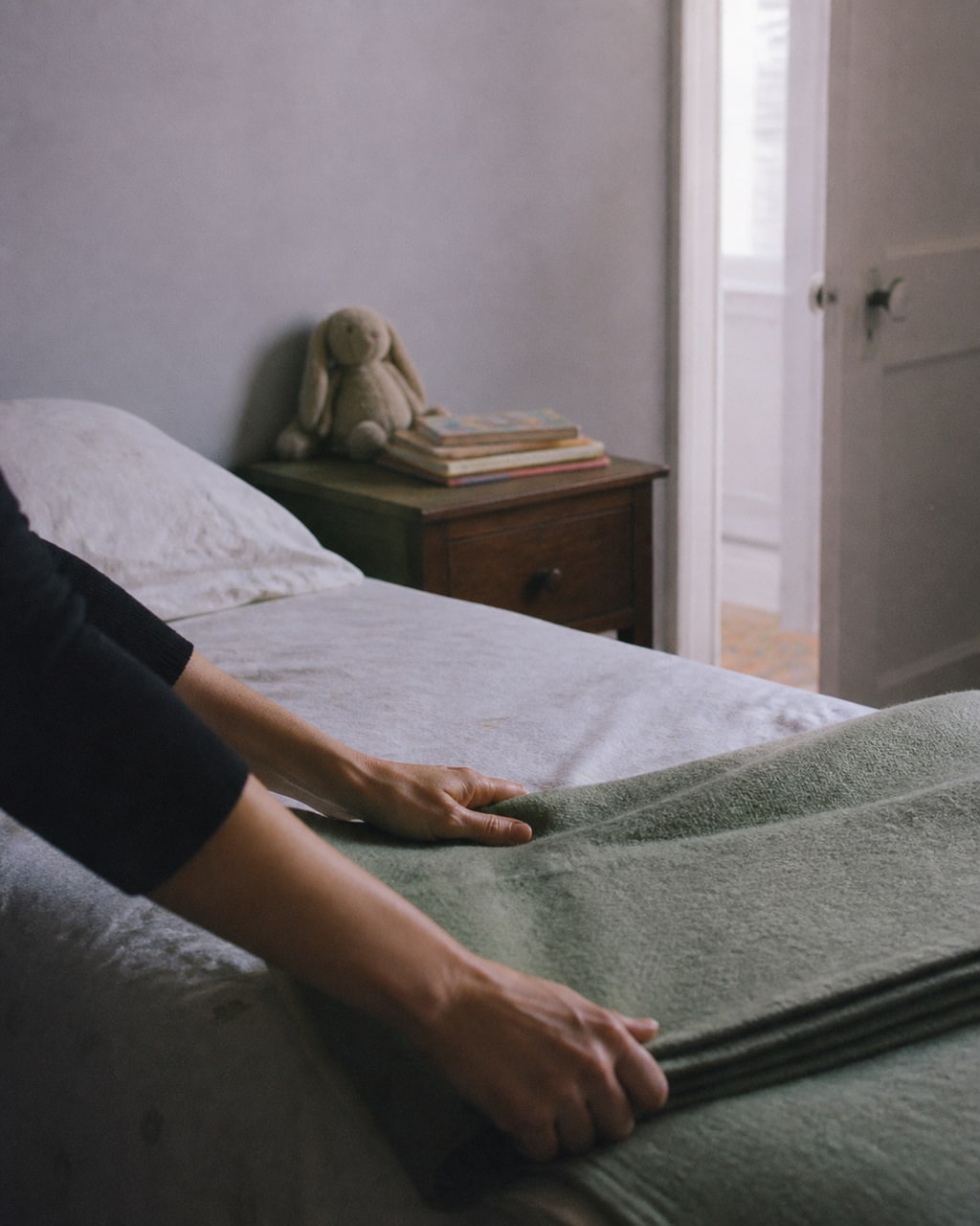 A parent's hands smoothing a child's made bed in a small, bright bedroom on a quiet morning