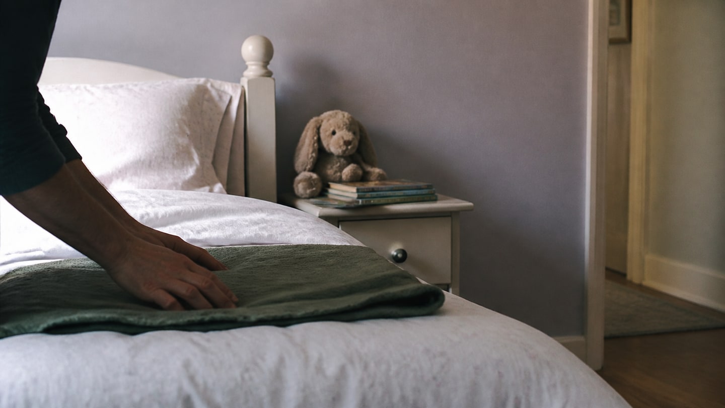 A parent's hands smoothing a child's made bed in a small, bright bedroom on a quiet morning